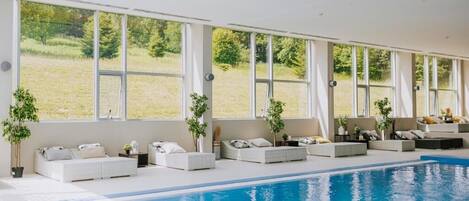 Piscina interior con tumbonas blancas y grandes ventanales con vistas a un campo de césped.