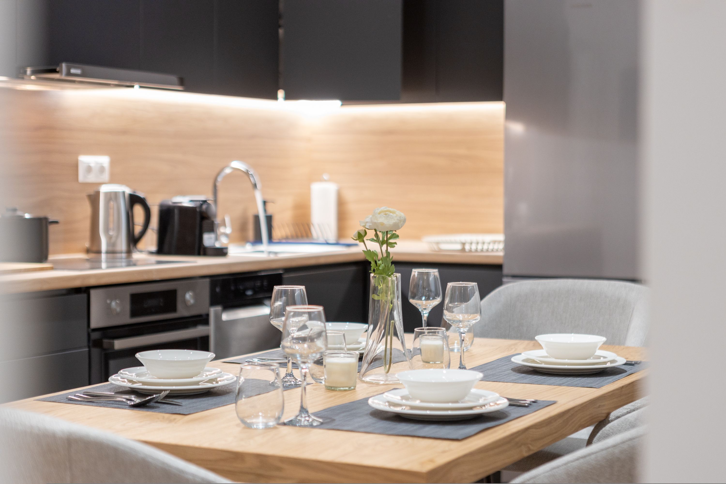 A modern dining area with a wooden table set for two, equipped with plates, glasses, and a vase with a flower.