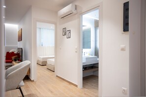 Een moderne hotelkamer met een houten vloer, een grijze fauteuil, een rode bank, een wit bed, een badkamer met spiegel en wastafel, en twee kunstwerken in lijsten aan de muur.