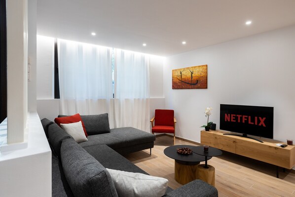 A modern living room with a grey sectional sofa, a red chair, a wooden TV stand, and a large flat-screen TV displaying "NETFLIX".