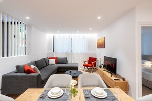 Een moderne woonkamer met een hoekbank, een eettafel gedekt voor twee personen, een televisie en een gezellige zithoek met een rode fauteuil.