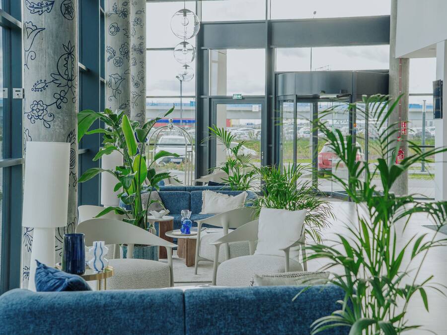 A modern hotel room with a large window offering a view of the airport. The room features a blue sofa, white armchairs, and several potted plants.