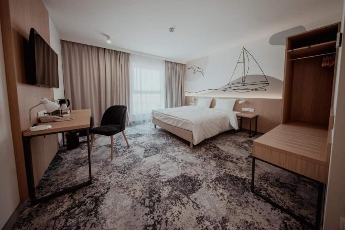 A hotel room with a bed, a desk with a chair, a TV, a wardrobe, and a large window with curtains.