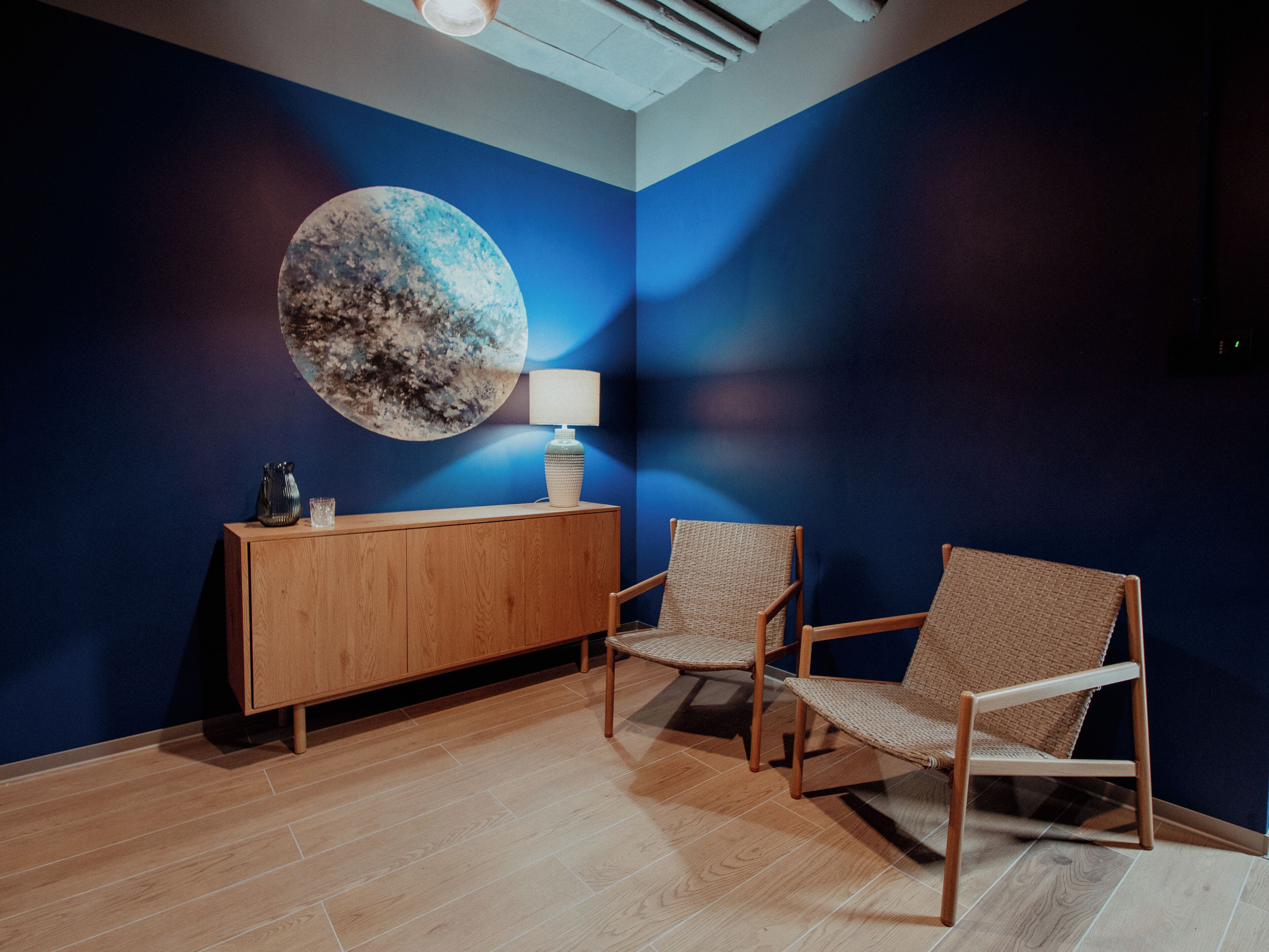 A room with a wooden cabinet, two wicker chairs, a large wall mural, and a floor lamp.