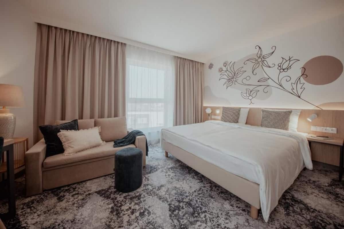 A modern hotel room with a large bed, a sofa, a small ottoman, and a decorative wall piece.