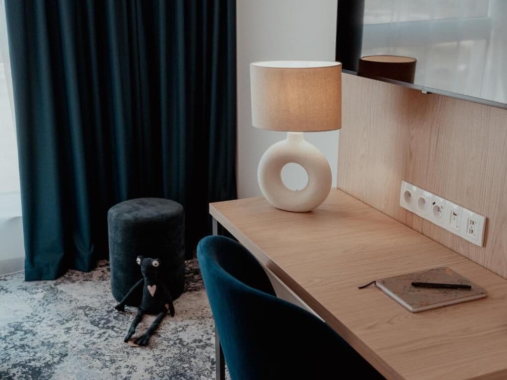 A modern room with a wooden desk, a lamp, a small ottoman, and a window with curtains.