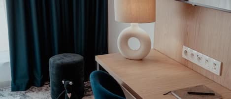 A modern room with a wooden desk, a lamp, a small ottoman, and a window with curtains.