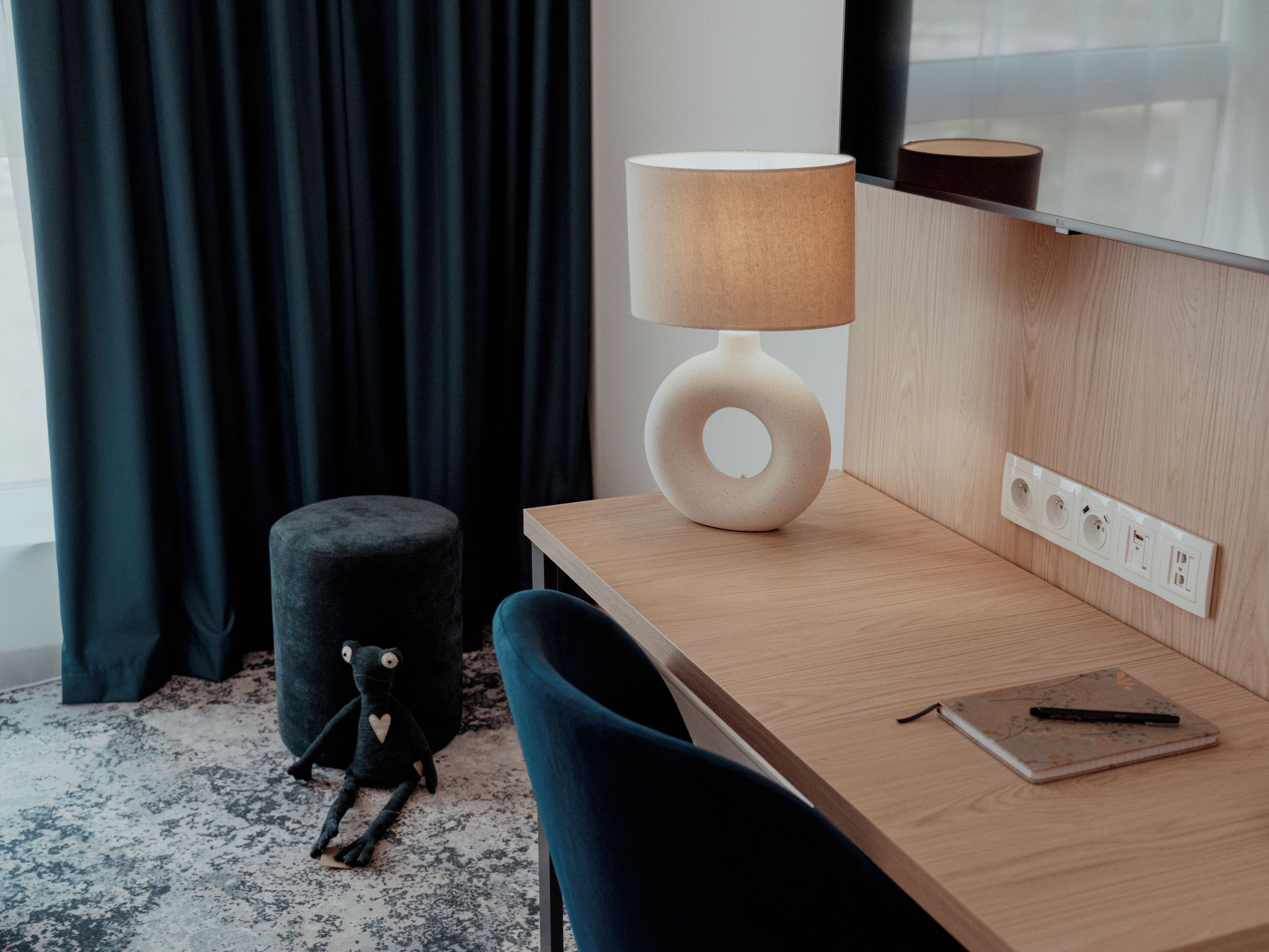 Una habitación de hotel moderna con un escritorio de madera, una lámpara, un otomano acolchado y una libreta con un bolígrafo.