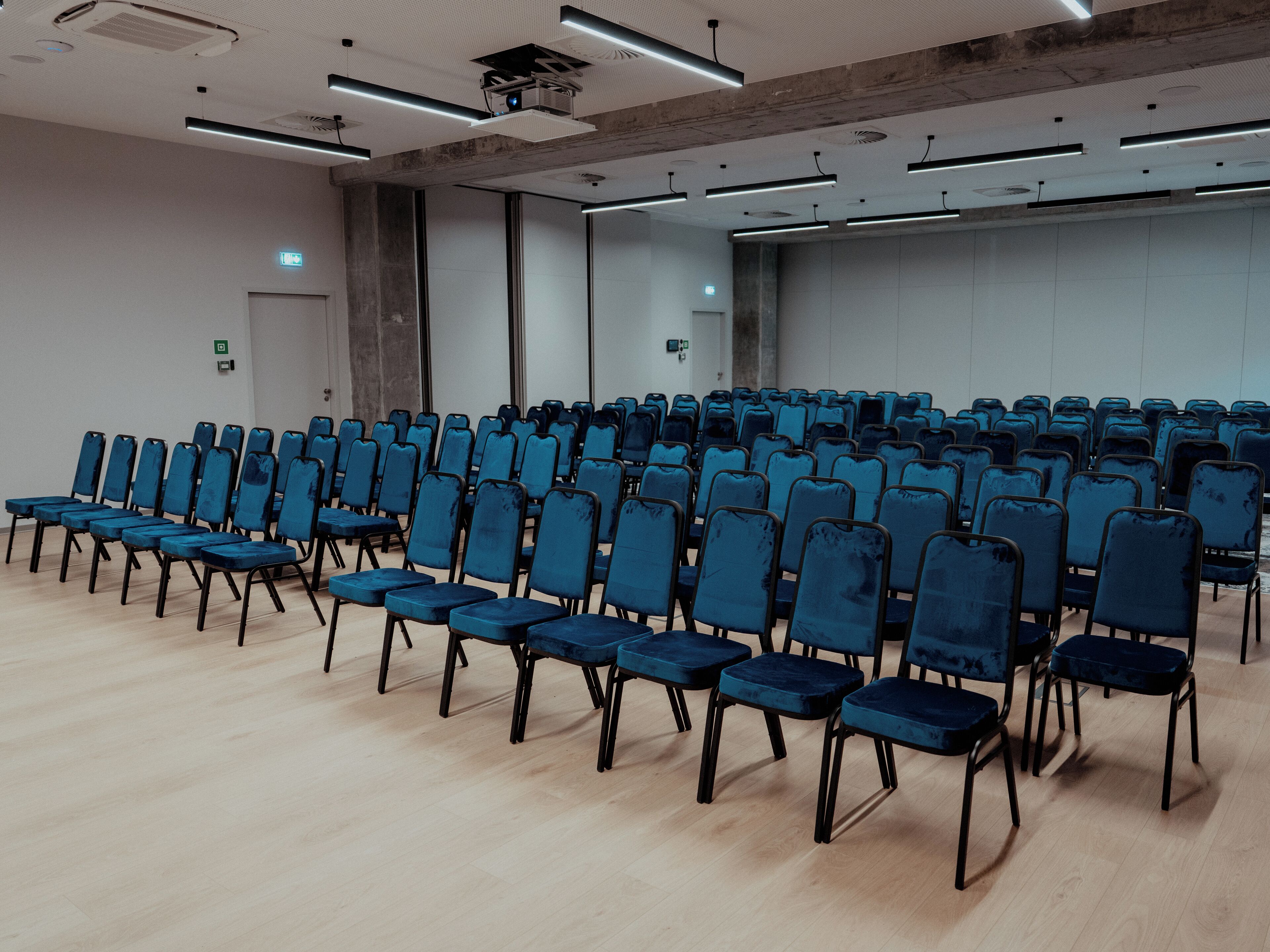 Uma sala de conferências com fileiras de cadeiras azuis, um projetor e um espelho grande na parede.