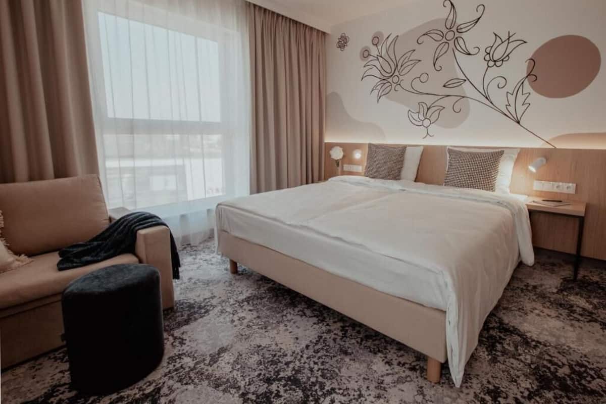 A hotel room with a large bed, a sofa, a small ottoman, a nightstand with a lamp, and a decorative wall mural.