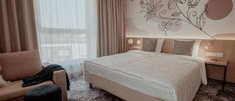 A hotel room with a large bed, a sofa, a small ottoman, a nightstand with a lamp, and a decorative wall mural.