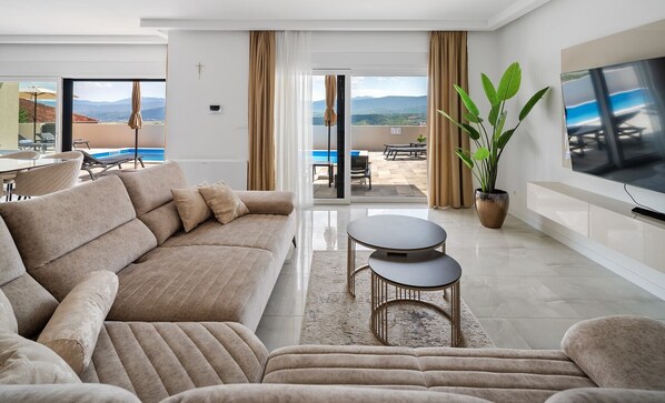 A modern living room with a beige sofa, two round tables, a large potted plant, and a flat-screen TV.