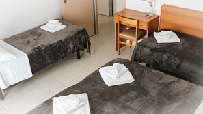 A hotel room with two beds, each with a grey blanket and white towels. There is a wooden desk with a chair, a small table with a vase of flowers, and a door. - Hotel Fattori (Cesenatico)