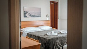 A neatly made bed with a grey blanket, a wooden headboard, and a white pillow. There is a wooden nightstand beside the bed with a folded white towel on it. A framed picture of a pier and waves is on the wall above the bed. - Hotel Fattori (Cesenatico)
