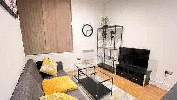 Uma sala de estar com um sofá cinzento, almofadas amarelas, uma mesa de centro de vidro, um aparador preto e uma planta numa prateleira.