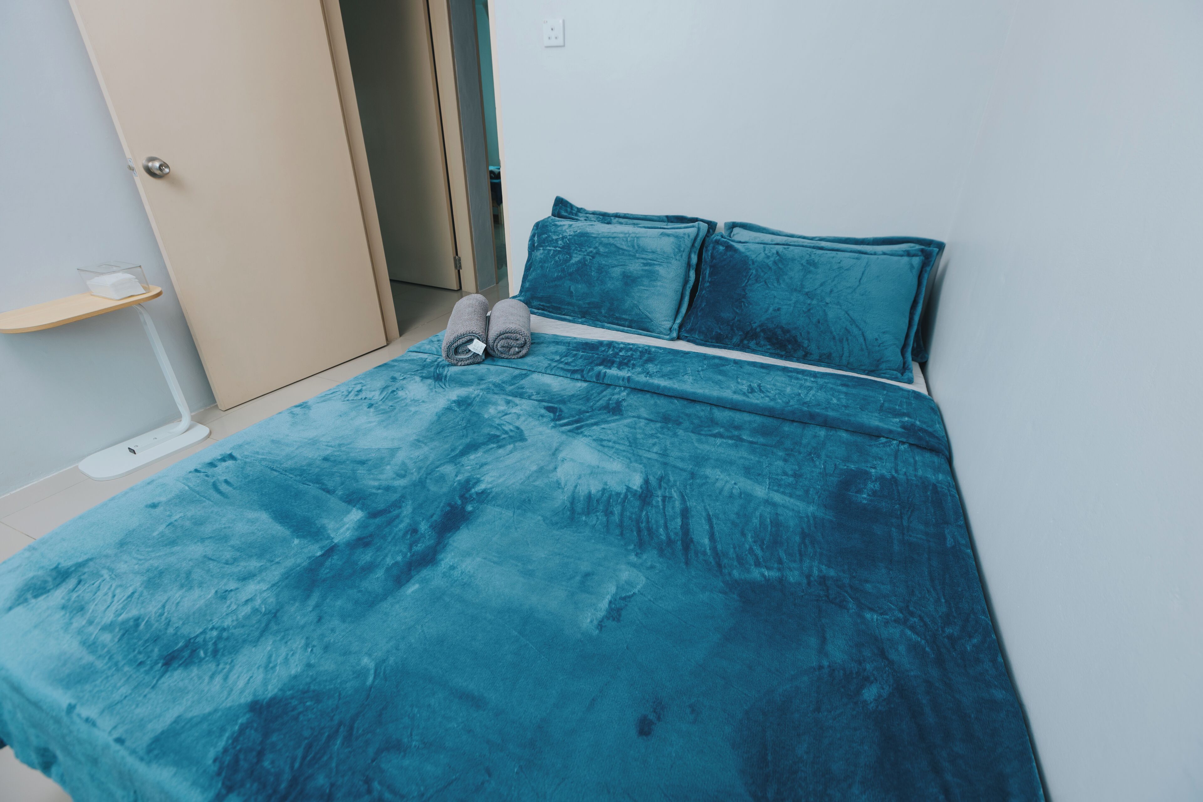 Un lit avec une couvre-lit et des oreillers de couleur turquoise, une table de chevet avec un récipient en verre, et une porte sur la gauche.