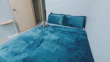 Un lit avec une couvre-lit et des oreillers de couleur turquoise, une table de chevet avec un récipient en verre, et une porte sur la gauche.