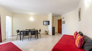 Ett kompakt bostadsutrymme med en matplats, en platt-TV, en soffa med färgglada kuddar och en dörr som leder till ett annat rum.
