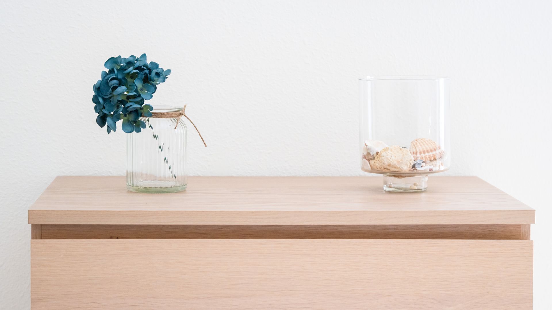 Two glass containers on a wooden surface; one holds blue flowers and the other seashells.