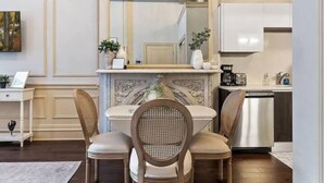 A dining area with a white table, two chairs, a decorative mirror above, and a kitchen area with a dishwasher.