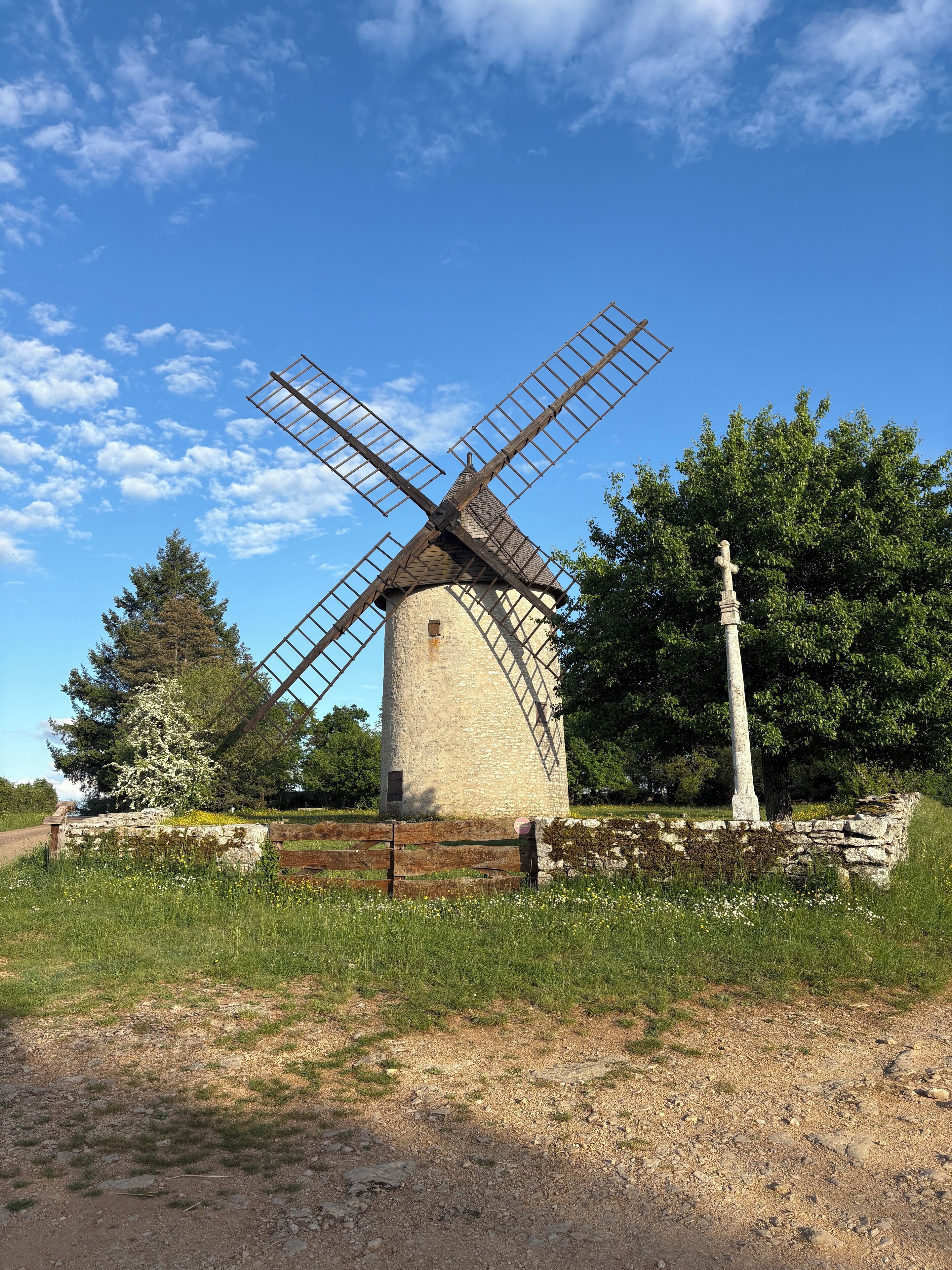 A traditional windmill with a stone base and wooden windmill blades, surrounded by greenery and a clear sky.