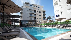 A swimming pool with sun loungers and umbrellas, surrounded by a multi-story building with balconies.