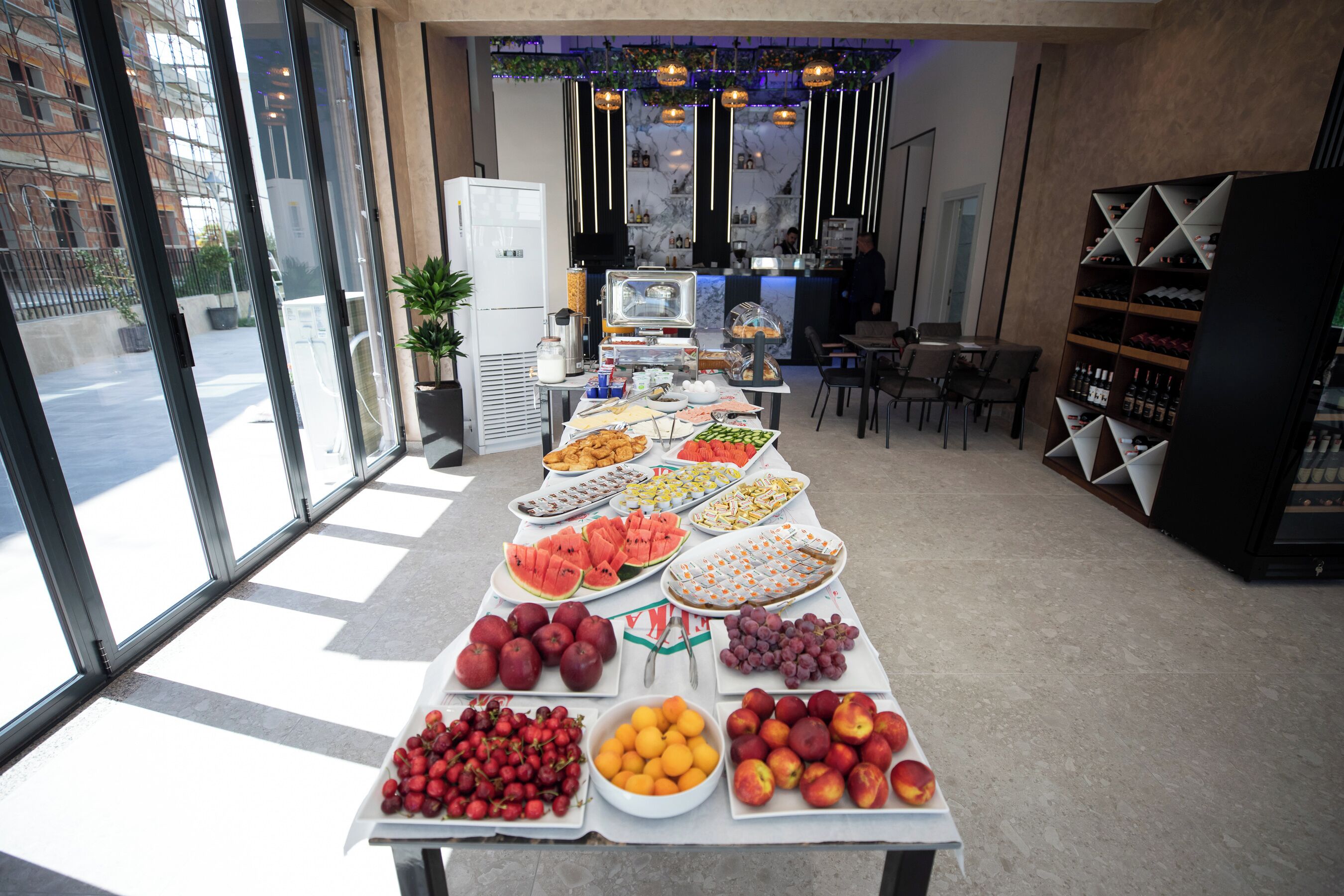 Una mesa de buffet con diversas frutas y aperitivos, un refrigerador y una zona de bar con estantes llenos de botellas.