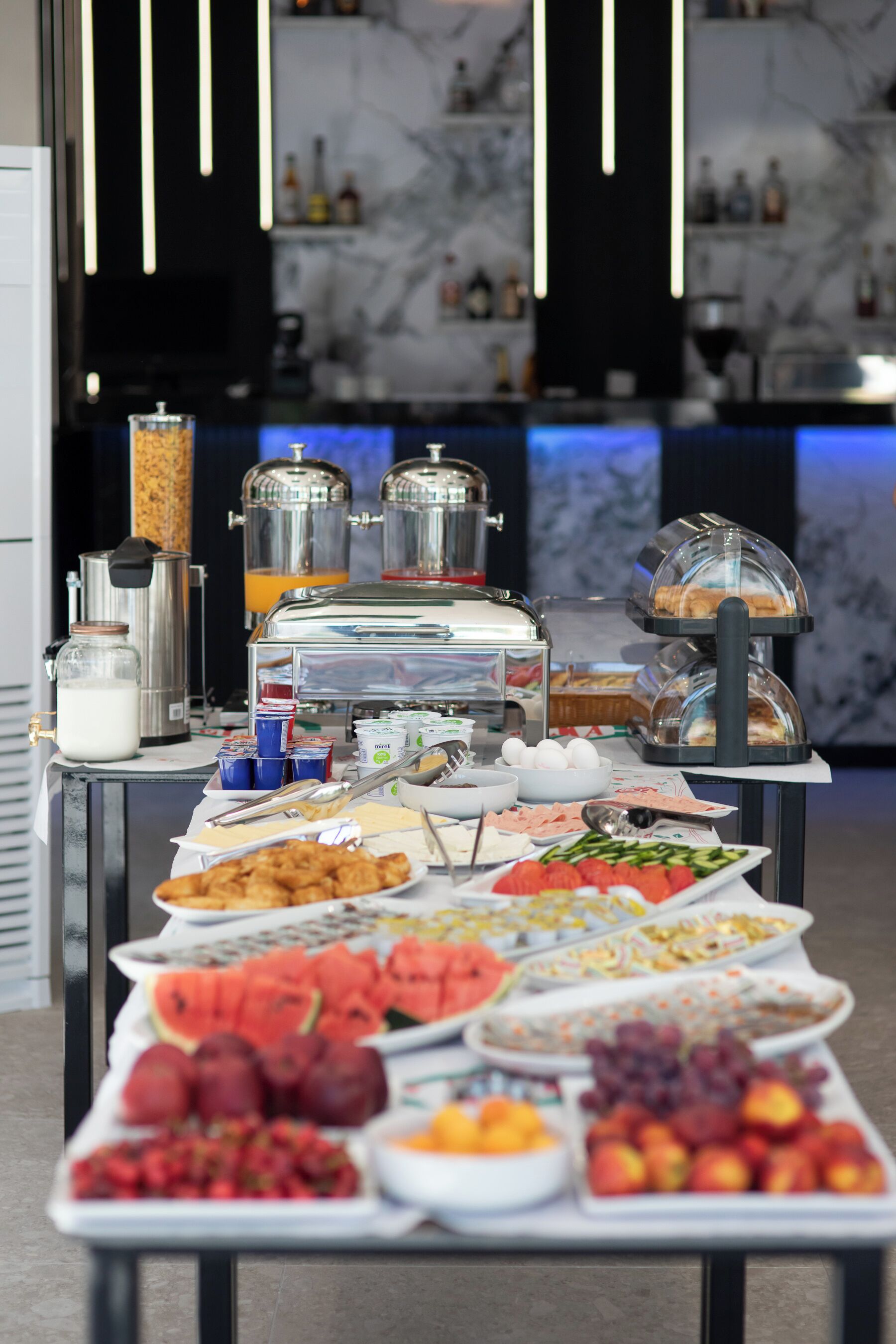 Tarjolla on runsas buffet, jossa on monenlaisia ruokia ja juomia, kuten hedelmiä, leivonnaisia ja meijerituotteita. Ne on aseteltu kauniisti pöydälle, ja taustalla näkyy moderni keittiö.