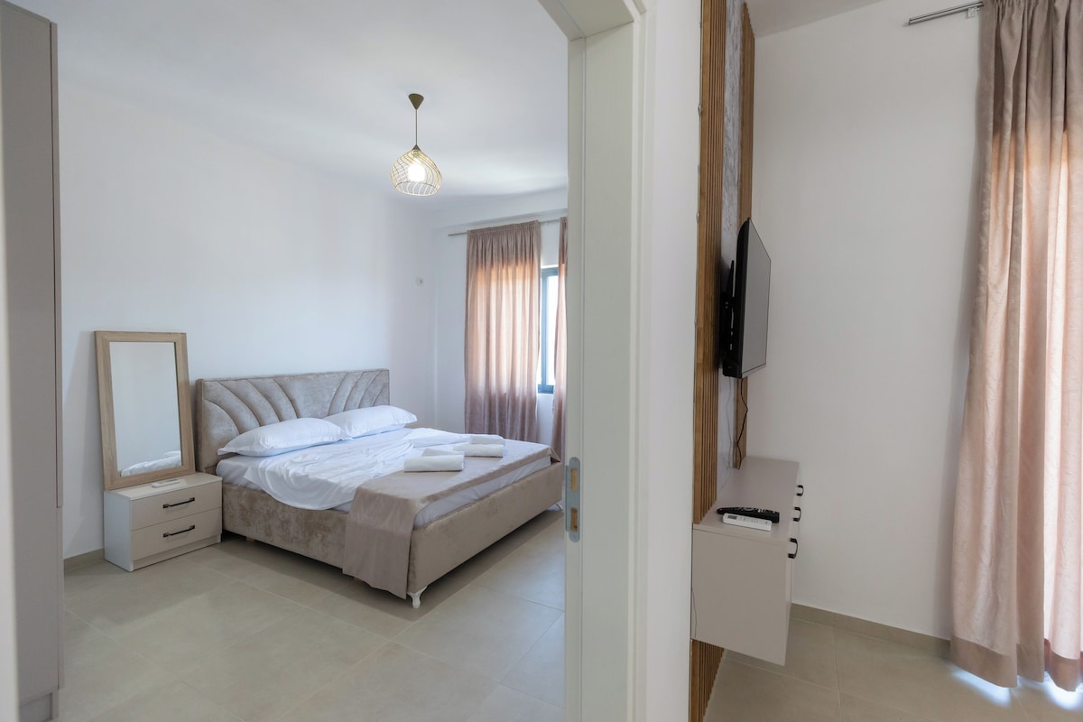 A bedroom with a bed, bedside table, mirror, TV, and curtains.