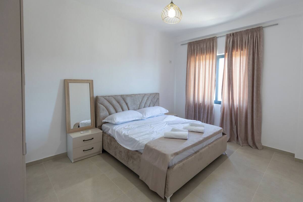 A neatly arranged bedroom with a bed, a bedside table, a mirror, and a window with curtains.