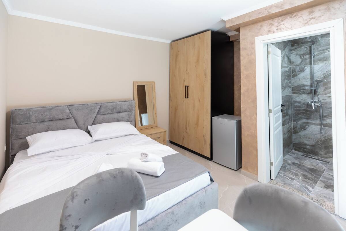 A modern bedroom with a bed, a wardrobe, a small fridge, and a shower area.