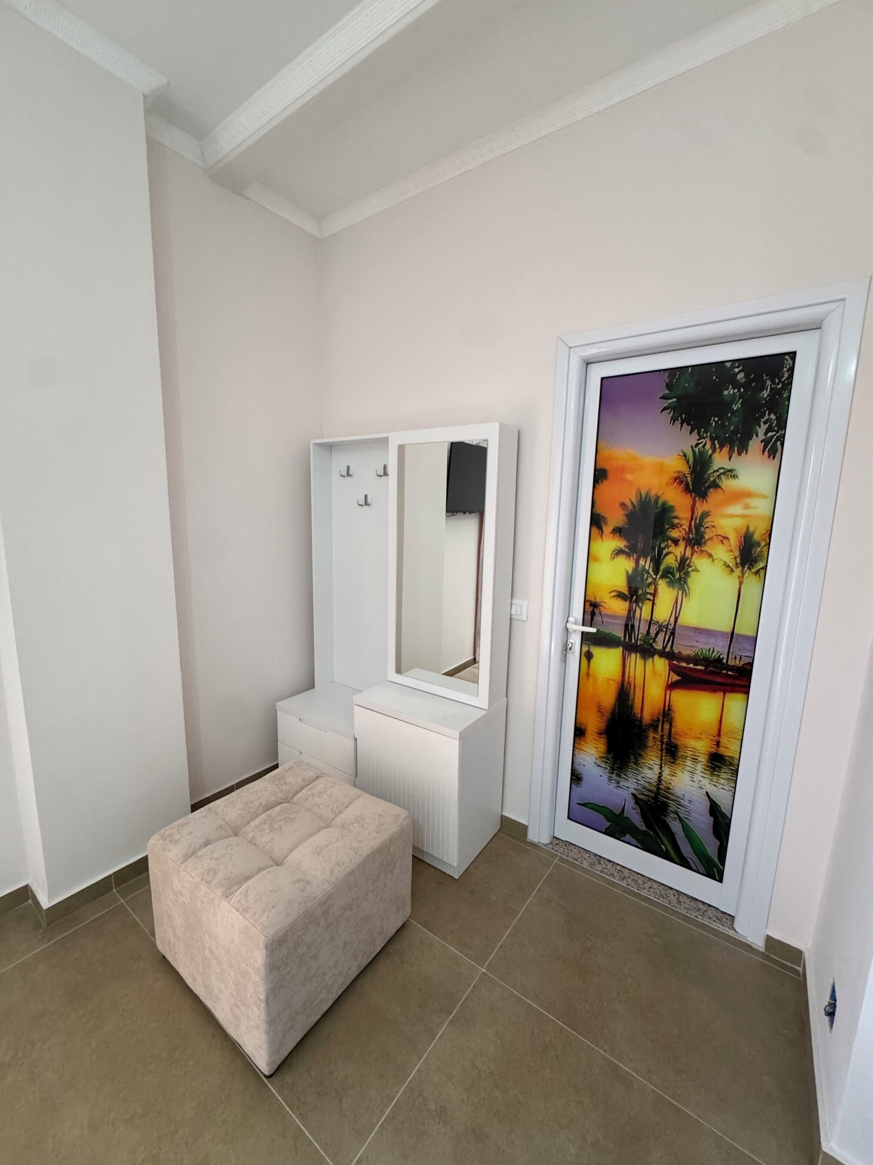 Una habitación con un tocador blanco, un reposapiés beige y una puerta con una pintura de atardecer.