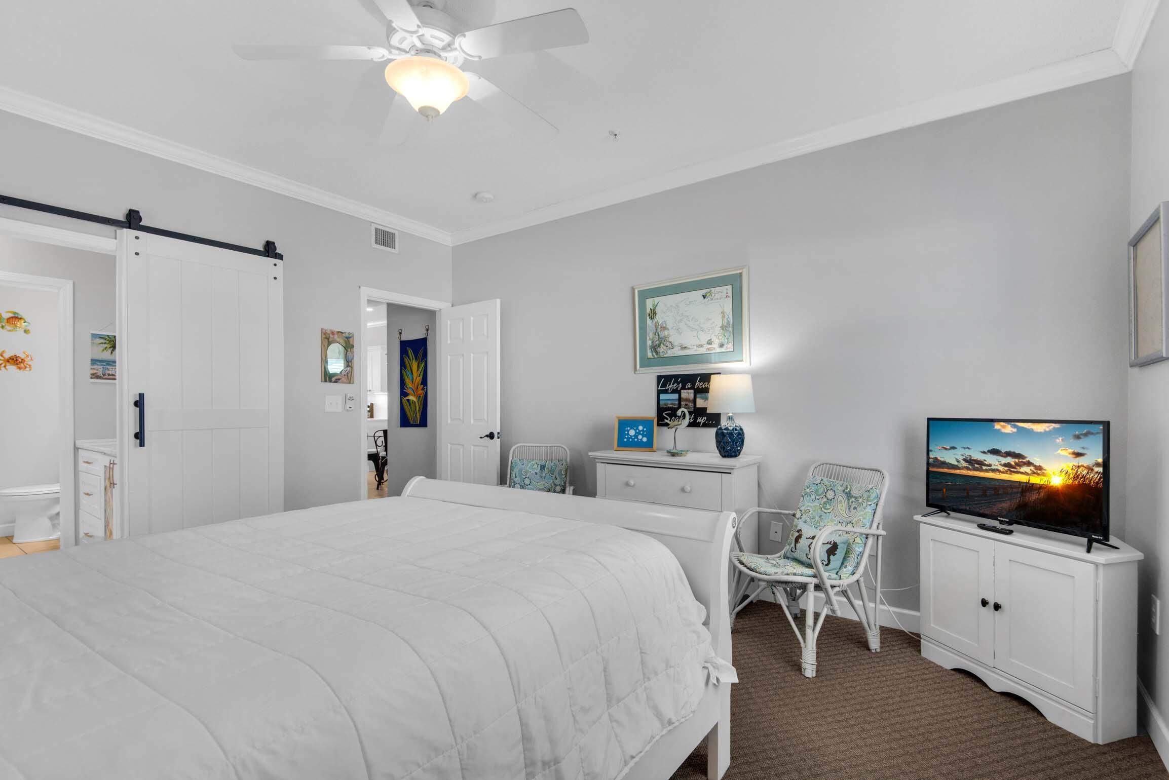 Un dormitorio con una cama, televisión, una silla, escritorio y ventilador de techo.