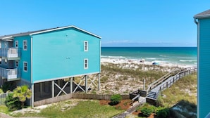 Un bâtiment turquoise à deux étages, doté d’un balcon et surélevé sur des piliers, qui surplombe une plage avec une promenade en bois.