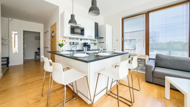 A modern kitchen with a central island, white bar stools, and a grey sofa in a bright living area with large windows. - Apartamenty Sun & Snow Golden Tulip (Miedzyzdroje)