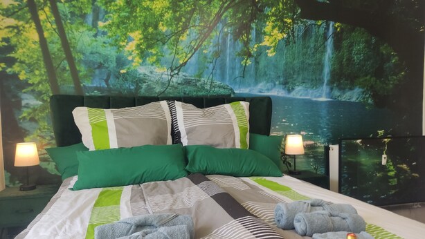 A bedroom with a bed, green and white bedding, two bedside lamps, and a forest-themed wall mural. - Apartment Classic (Jülich)
