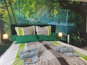 A bedroom with a bed, green and white bedding, two bedside lamps, and a forest-themed wall mural. - Apartment Classic (Jülich)