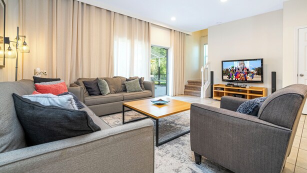 A modern living room with a sofa, armchair, and coffee table, a flat-screen TV, and a wooden entertainment unit. - Family-Friendly 3BR Townhome at Encantada Resort ● Pool, BBQ & Near Disney! (Kissimmee)