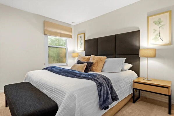 A bedroom with a large bed, a bedside table, a bench, a window with a view, and a wall-mounted lamp.
