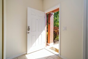 A white door with a doormat and a small outdoor area with a brick pillar and greenery.
