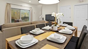 A dining area with a long table set for a meal, a sofa in the background, and a television on the wall. - Family-Friendly 3BR Townhome at Encantada Resort ● Pool, BBQ & Near Disney! (Kissimmee)