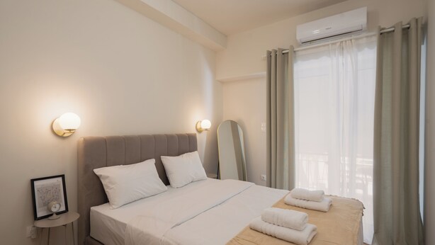 A modern hotel room with a large bed, a side table with a clock, and a view of the outdoors through sheer curtains. - "Acropolis Kallisti" 2BR/2BA near Metro (Athina)