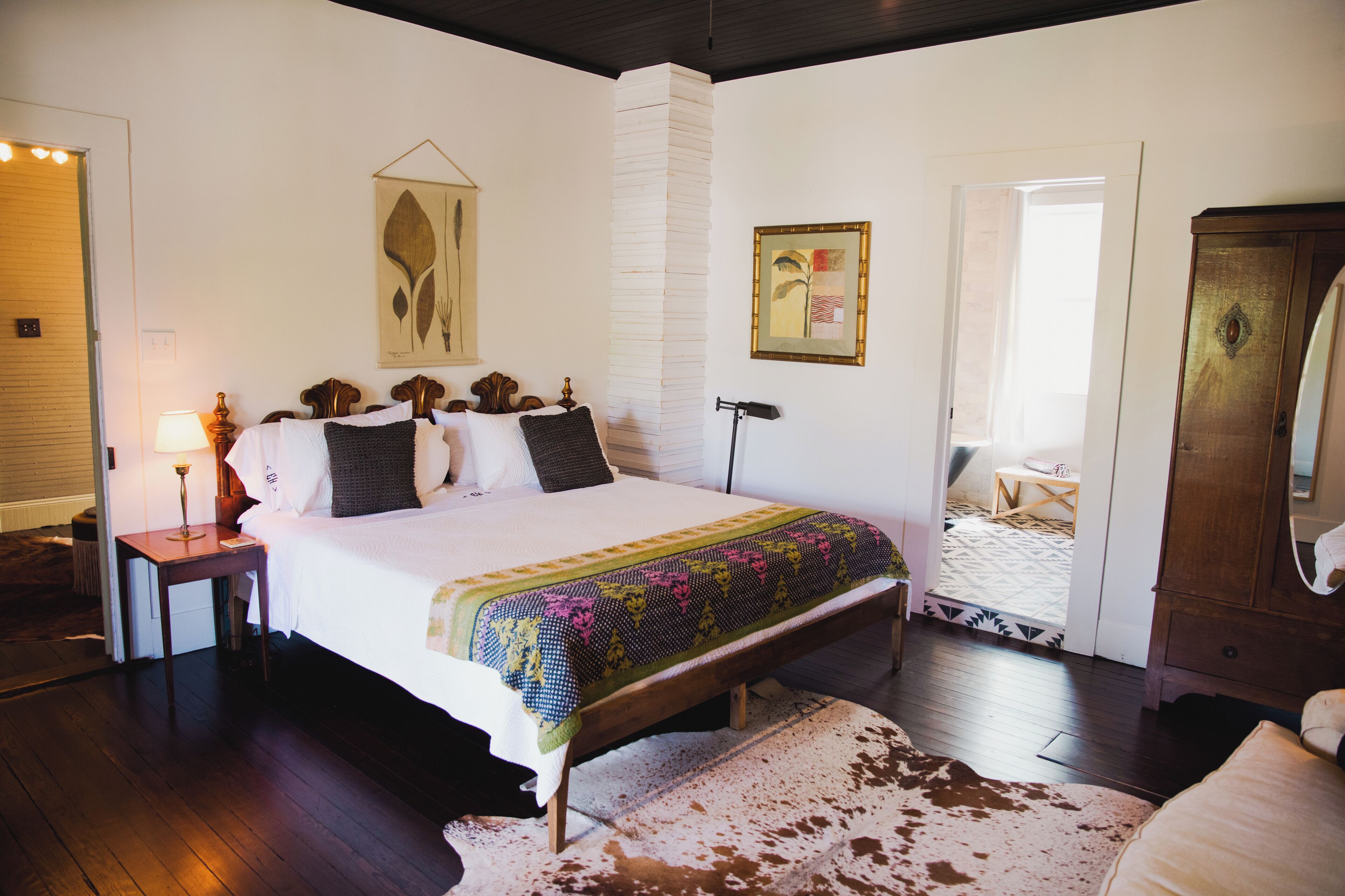 Une chambre avec un grand lit, une table de chevet en bois, une lampe, une estampe botanique encadrée et une porte donnant sur une autre pièce.
