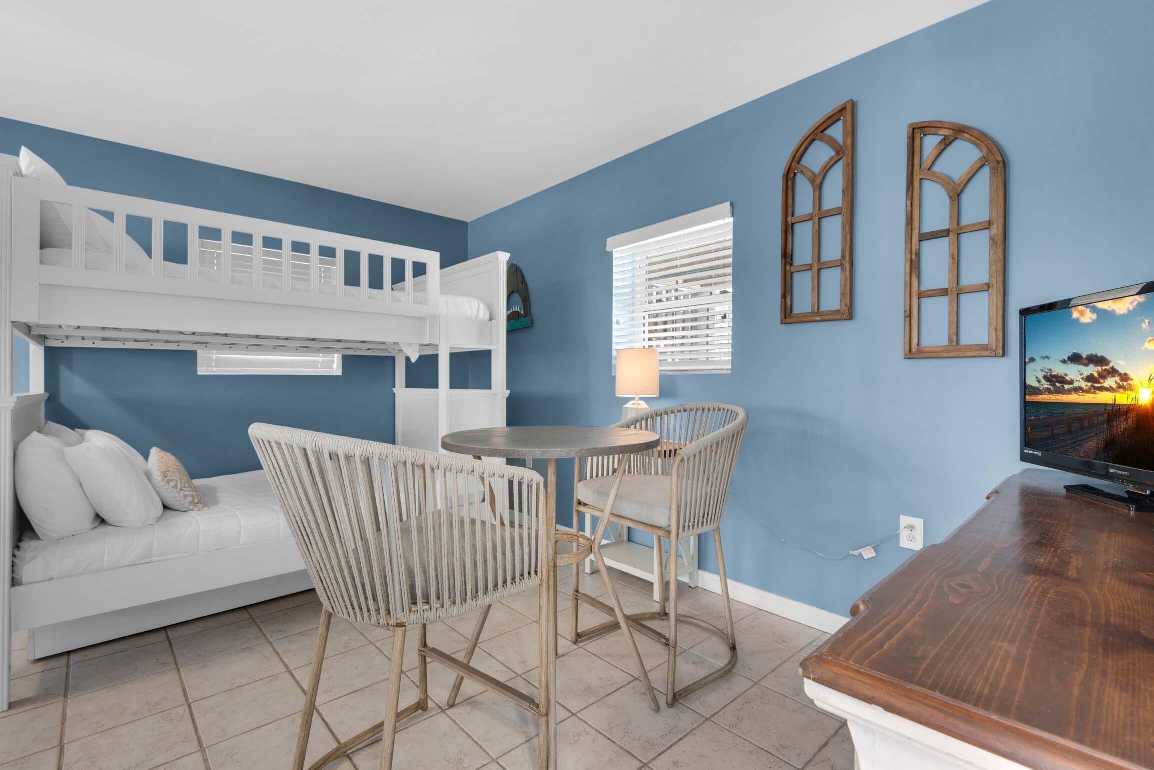 A room with a bunk bed, a white sofa, two wicker chairs, a small round table, a wooden desk, and a TV on a wooden stand.