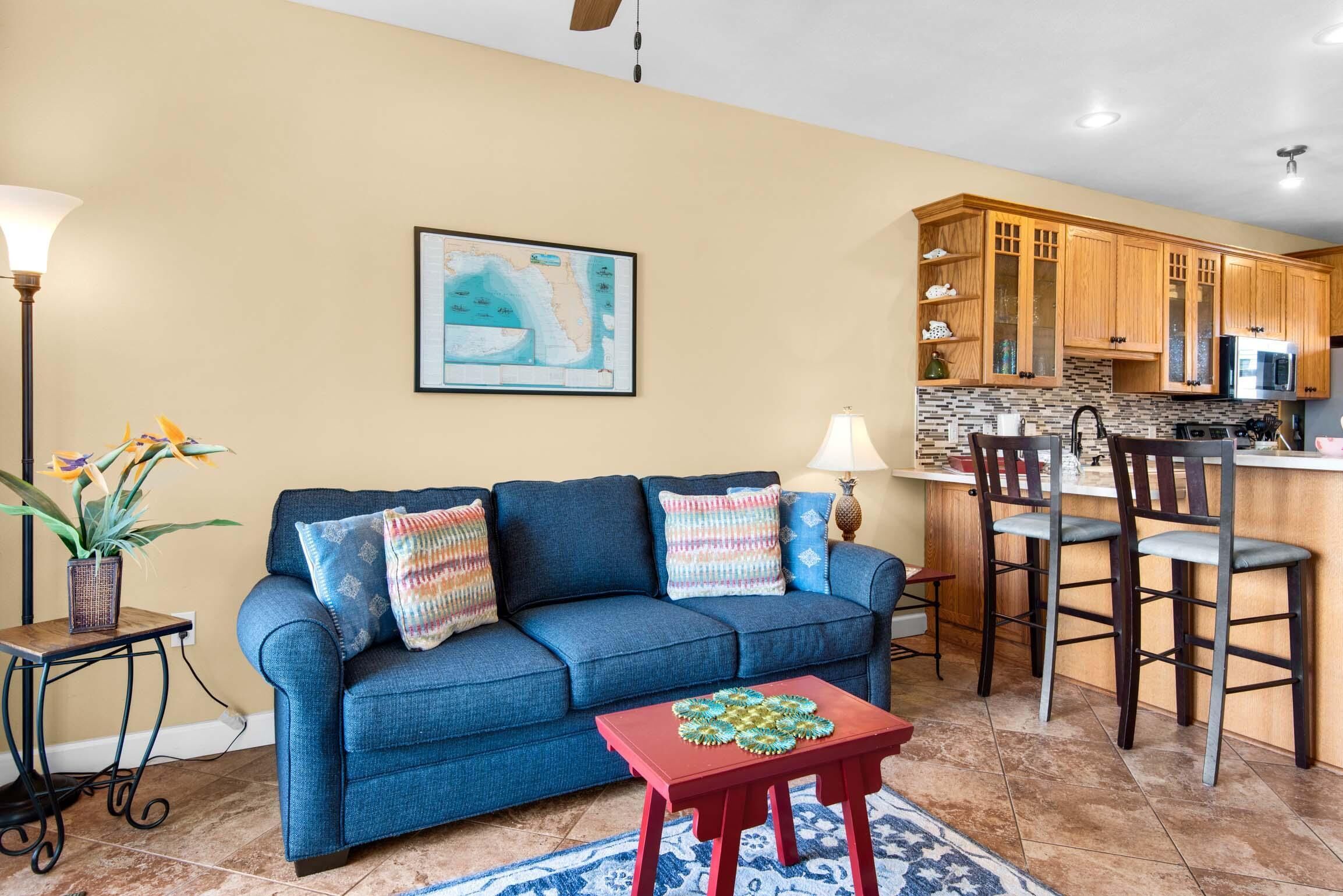 A living room with a blue sofa, a red coffee table, and a kitchen area with wooden cabinets and a tiled backsplash.