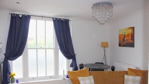 A living room with a sofa, curtains, a chandelier, a painting, and a table with chairs. - Stylish & Spacious 1 bed flat next to station (London)