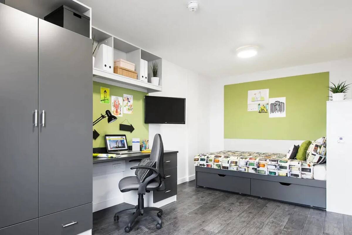 Une chambre moderne avec une armoire grise, un bureau avec un ordinateur, un fauteuil de bureau noir, un lit avec une couvre-lit à motifs et des murs verts.
