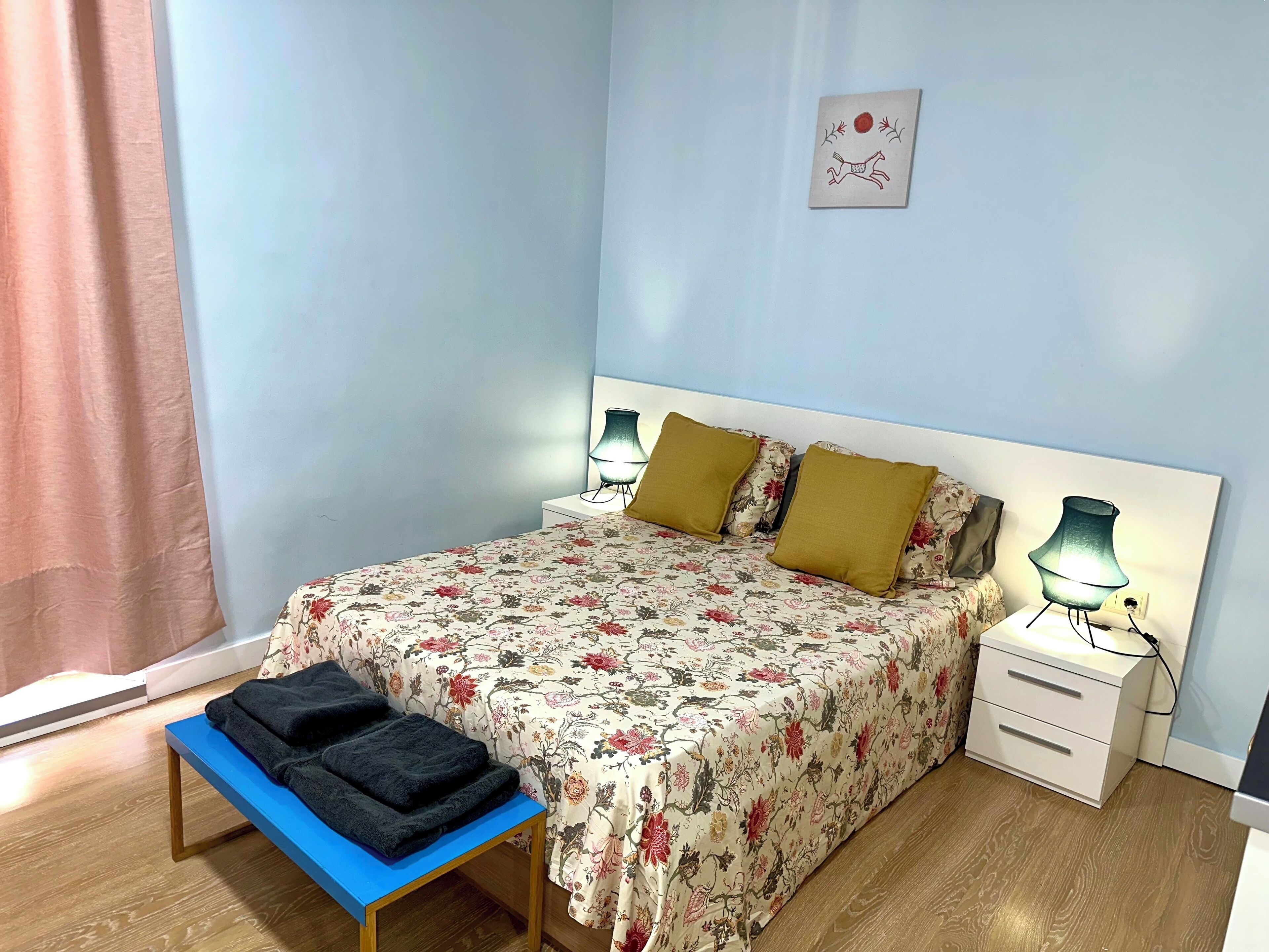 A bedroom with a floral bedspread, two pillows, a bedside table with lamps, a bench with a folded blanket, and a wall decoration.