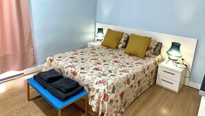 Ein Schlafzimmer mit floralem Bettbezug, zwei Kissen, einem Nachttisch mit Lampen, einer Bank mit einer zusammenfaltbaren Decke und einer Wanddekoration.