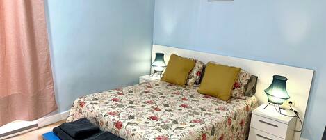 A bedroom with a floral bedspread, two pillows, a bedside table with lamps, a bench with a folded blanket, and a wall decoration.